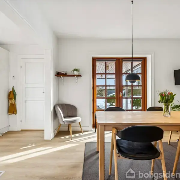 Et spisebord med sorte stole står i et lyst rum ved en glasdør. Over bordet hænger to lamper. På bordet er der en vase med blomster, og væggen har en fladskærm. En stol og jakker er ved siden af en hvid dør.