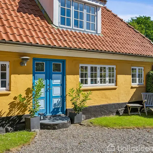 Gult hus med blå hoveddør og rød tag, omkranset af grøn hæk. To stole står på grus foran huset. Solrig dag med blå himmel.