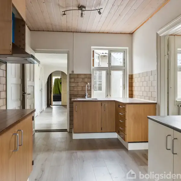 Køkken med træloft og flisevægge. Træskabe langs væggene. Rummets åbne design viser en vask ved vinduet. Dagslys strømmer ind gennem vinduerne.
