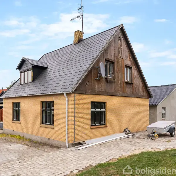 Hus med gule mursten og spidst sort tag står på brolagt grund. Omgivet af bygninger, græsplæne og træer. Trailer og byggematerialer ses ved siden af huset.