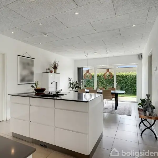 Køkkenø med vask står centralt i et lyst køkken. I baggrunden er der spisebord med stole og store vinduer, der giver udsigt til en grøn have.