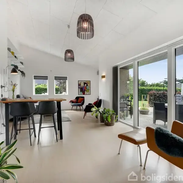 En moderne stue med stilfuld indretning, hvor to sorte lamper hænger fra loftet. Store vinduer åbner ud til en terrasse med haveudsigt. Grønne planter pynter rummet.