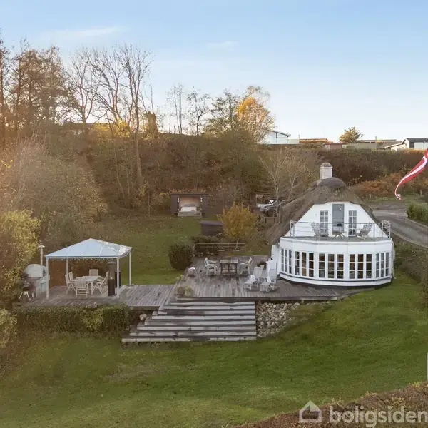 Et rundt, hvidt hus med store vinduer ligger i en frodig have med en terrasse. En pavillon med havemøbler står til venstre for huset. En flagstang bærer et flag.