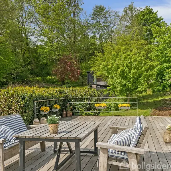Terrassemøbler står på en træterrasse med stribede hynder, omringet af en frodig have med grønne træer og buske. Blomster i krukker pynter omgivelserne under en klar blå himmel.