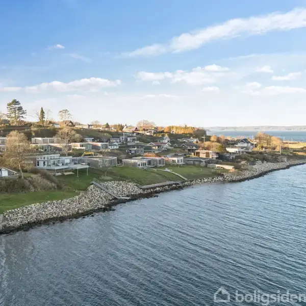 Kystnært boligområde langs vandet. Huse ligger tæt ved kystlinjen med en grøn skråning, mens havet strækker sig i baggrunden under en klar, blå himmel.
