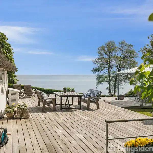 Stråtagshus med stor træterrasse, har havemøbler opstillet med udsigt over havet. Store træer og grønne planter omkranser området. Klar blå himmel i baggrunden.