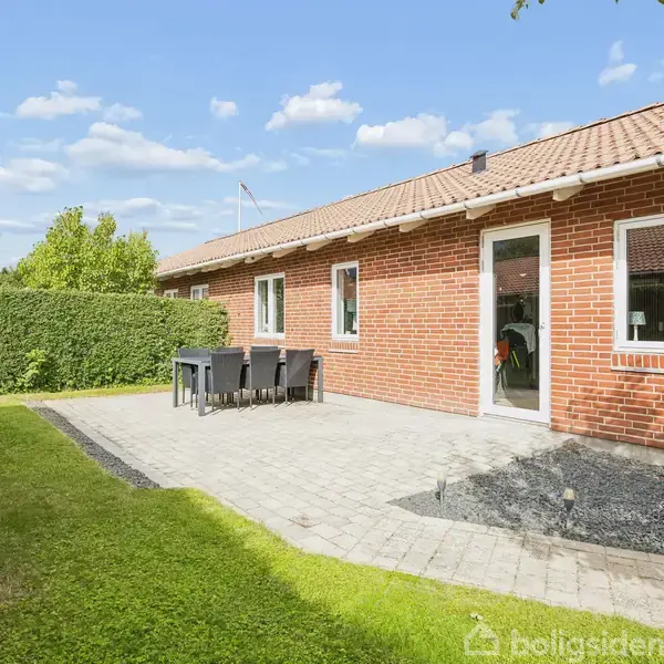 Rødt murstenshus med en fliseterrasse, hvor et bord med stole står. Omgivet af grøn græsplæne og hæk. Klar blå himmel med få skyer i baggrunden.