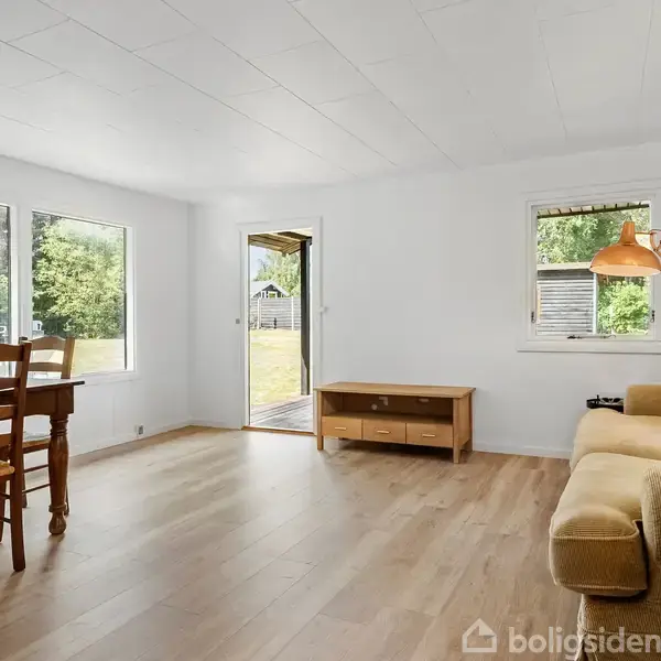 Stue med træbord og to stole ved store vinduer, cremefarvet sofa med en orange lampe ved siden af et lille vindue; dør åbner ud til have.