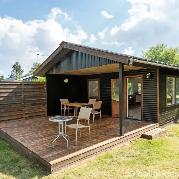 Sort træhus med åben terrasse indrettet med stole og borde, omgivet af grønne græsplæner og træer. Sollys skinner på terrassen og huset.