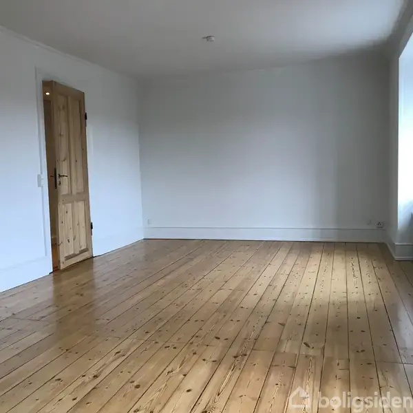 Et tomt rum med trægulv og hvide vægge. En naturlig trædør står på den ene side, og en stor vindueskarm lader lys skinne ind fra den anden side.