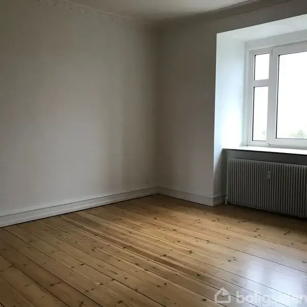 Et tomt værelse med trægulv og hvidmalede vægge. Et vindue med radiator nedenunder tillader lys ind. Rummet er enkelt og uden møbler.
