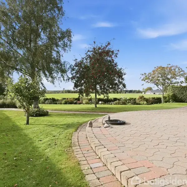 En flisebelagt terrasse står stille i en frodig have omkranset af grønne træer og hække. I baggrunden ses åbne græsarealer under en klar blå himmel.