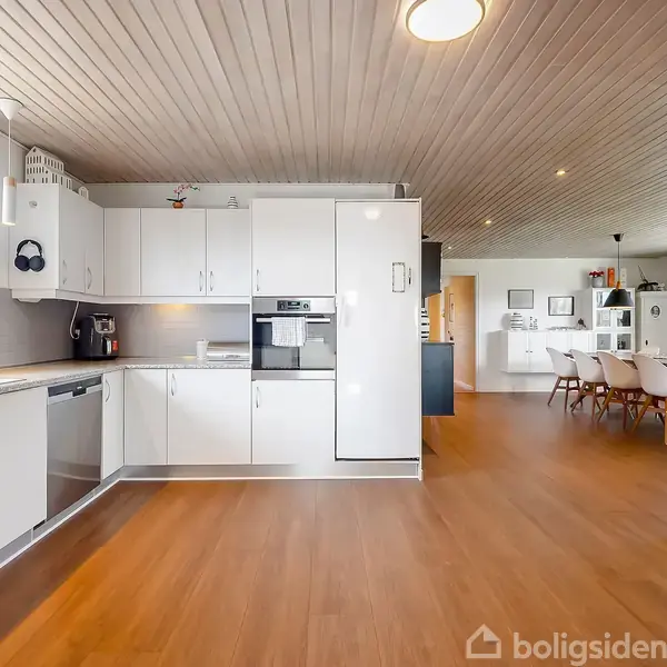 Et hvidt moderne køkken med indbyggede apparater og trægulv. På højre side ses en spiseafdeling med et bord og stole. Lyse vægge og loft giver rummet et åbent udseende.