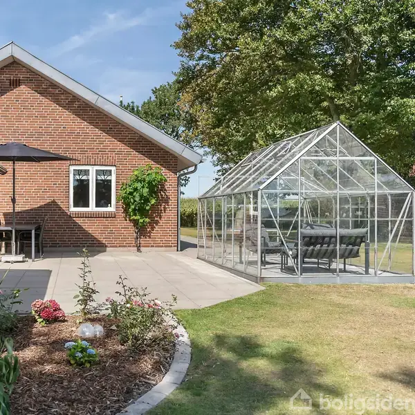 Rødt murstenshus med terrasse, hvor der står et bord og parasol. Ved siden af er der et drivhus med møbler. Haven er velplejet med blomsterbede og træer i baggrunden.