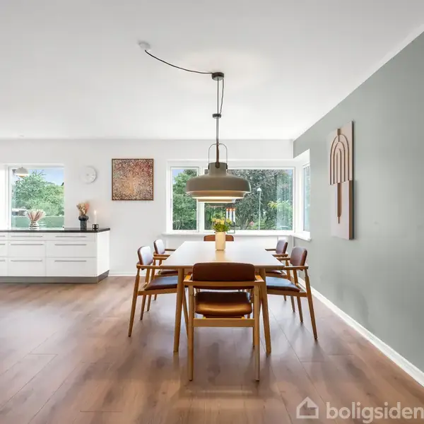 Spisebord i træ med seks stole, står centralt på et lyst trægulv. Over hænger en lampe. Til venstre er et moderne køkken med store vinduer, der lukker lys ind.