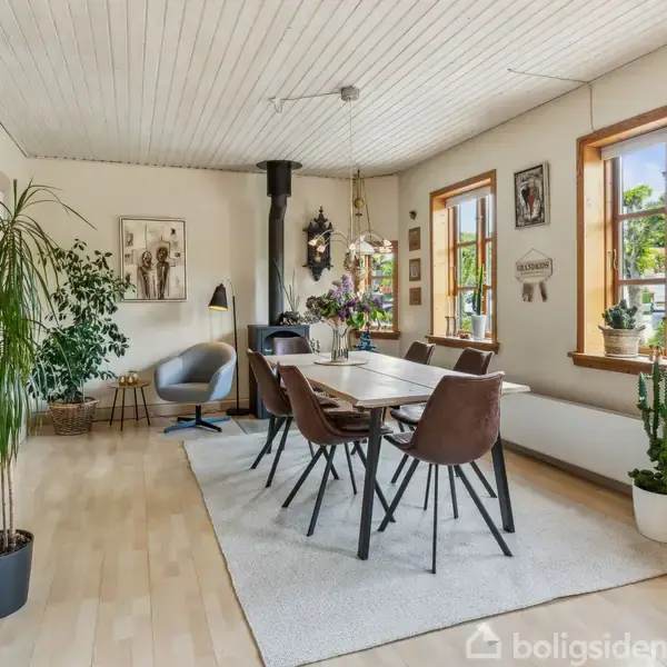 Spisebord står dækket med blomster, omgivet af seks stole i en lys stue. Væggene har kunstværker, planter, og en brændeovn. Store vinduer giver udsigt til en have.