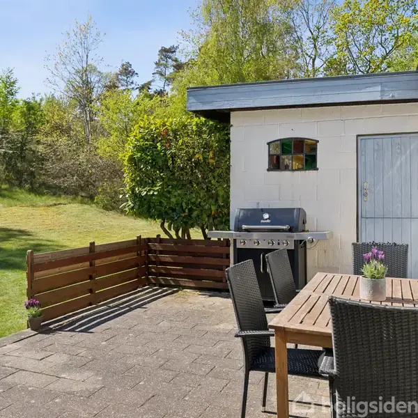 Terrassebord med fire stole står stille på flisebelagt terrasse. Grill ved murstensvæg. Baggrund har frodig have med græsplæne og træer under blå himmel.
