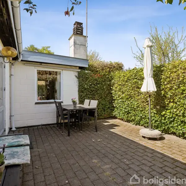 Terrasse med bord og stole omgivet af hække. En parasol står sammenfoldet ved siden af. Vægmonteret lampe og blomster på bænken under vinduet skaber en hyggelig atmosfære.