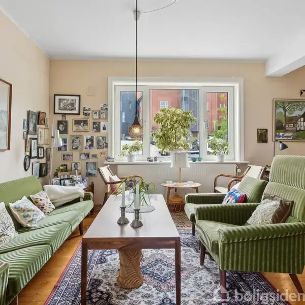 En stue med grønne møbler og et træbord, dekoreret med billeder og malerier på væggene. Et stort vindue slipper lys ind, og der er en plante på bordet.