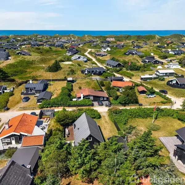 Mange huse ligger spredt i et kuperet, grønt landskab nær kysten. Husene er omgivet af græs og buske, mens havet ses i baggrunden under en klar blå himmel.