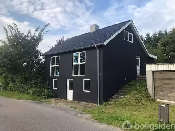 Et sortmalet hus med hvide vinduer står på en skråning. En græsbelagt bakke med trappe fører op til indgangen. Huset ligger ved siden af en vej og omgivet af træer.