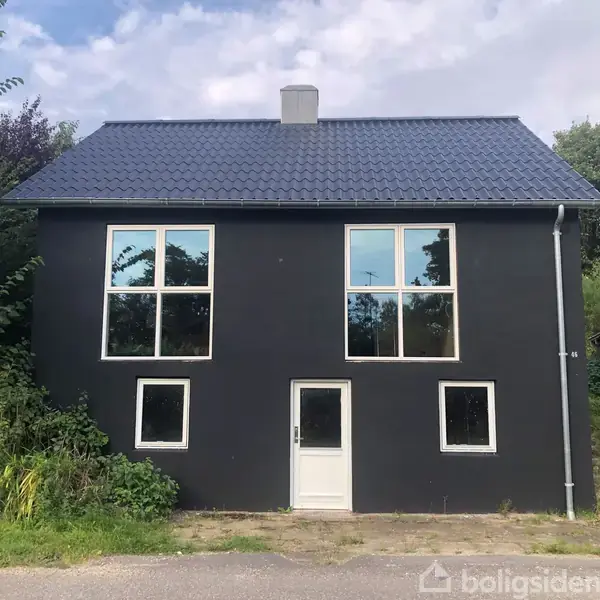 Sort hus med fire store vinduer og en hvid dør, omkranset af grønt buskads. Tørt betongulv foran og blågrå himmel med skyer i baggrunden.