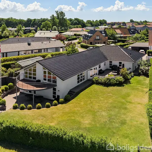 Hvide murstenshus med mørkt tag står i en velplejet have omgivet af hække og en flagstang. Huset har store vinduer og ligger i et beboelseskvarter med flere lignende huse.