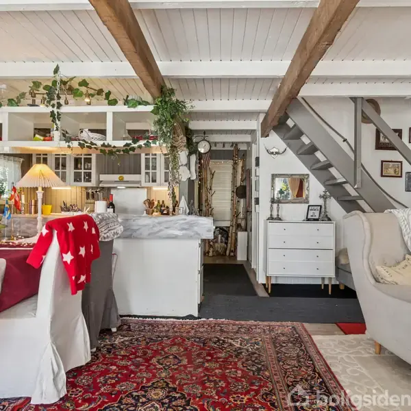 Stueområde med spisebord dækket af rødt og hvidt tæppe, omringet af stole og lænestol. Trappen fører til en øvre etage. Hylder og planter pryder køkkenet i baggrunden.