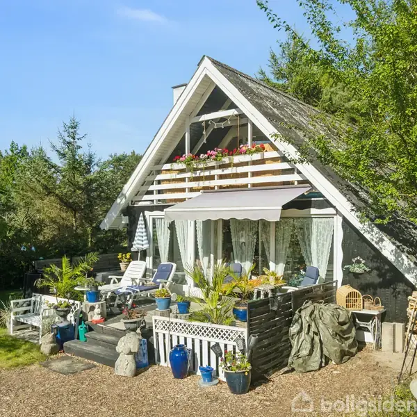 Et charmerende, sort-hvidt sommerhus med en terrasse fyldt med potteplanter og havemøbler. Huset ligger i en grøn have omkranset af træer og buske under en klar blå himmel.