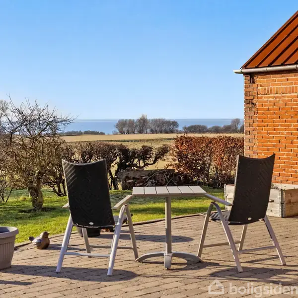 To stole og et bord står på en flisebelagt terrasse. I baggrunden ses en græsplæne, marker og hav med blå himmel. Ved siden af stolen er en krukke og nogle små figurer.