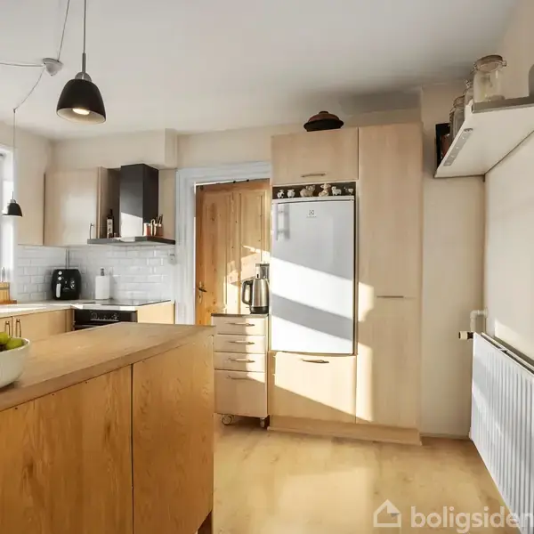 Træskab står i et lyst køkken med moderne indretning; en skål med frugt står på køkkenbordet. Vinduerne giver naturligt lys, og der er hvide fliser samt sort køkkenudstyr.