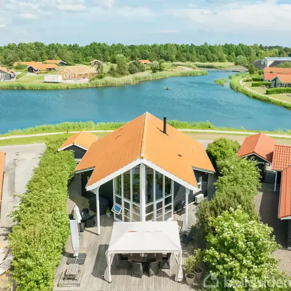 Hus med orange tag står ved en sø med flere omkringliggende huse og grøn bevoksning. Terrasse med pavillon er foran huset, omgivet af buske.