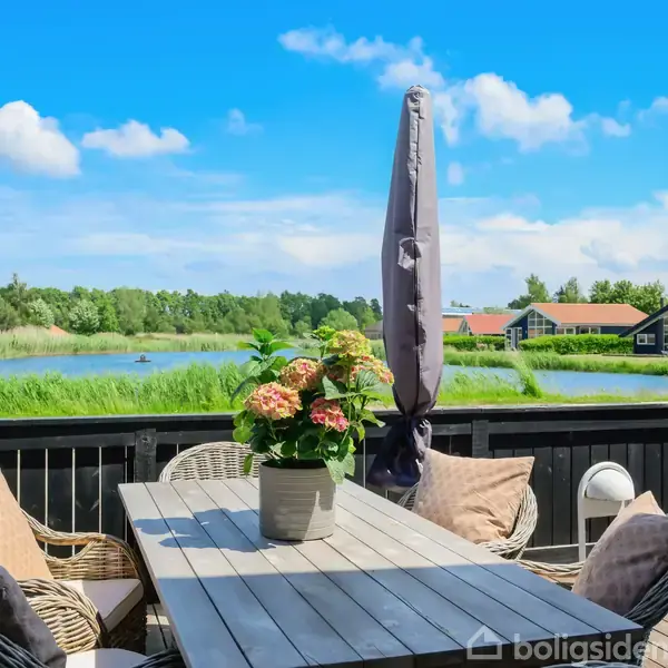 Et bord står på en terrasse med stole og en parasol. Udsigt over en sø med huse og natur i baggrunden under en blå himmel.