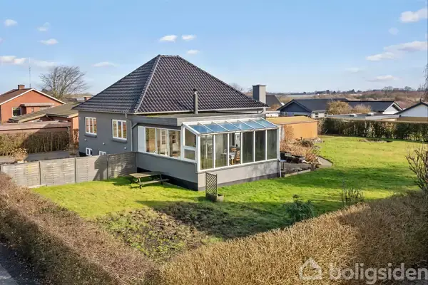 Hus med sort tag og udestue omringet af grøn have set fra bagsiden. Naboerne ses i baggrunden på en solrig dag.