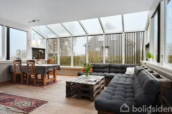 En lys stue med en sort sofa arrangeret i en L-form, står omkring et sofabord lavet af paller. Et spisebord med stole står ved siden af en glasvæg med udsigt til haven.