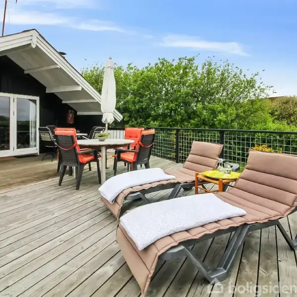 To solsenge med puder står på en træterrasse foran et sommerhus. En parasol og borde med stole er til stede, omgivet af grønne buske under en blå himmel.