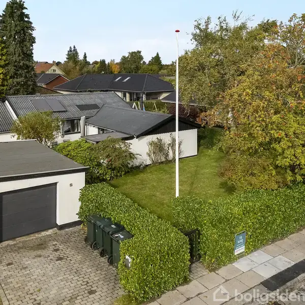 Et parcelhus med fladt tag står i en velplejet have med en flagstang. Huset er omgivet af grønne hække og store træer. Et skilt står foran.