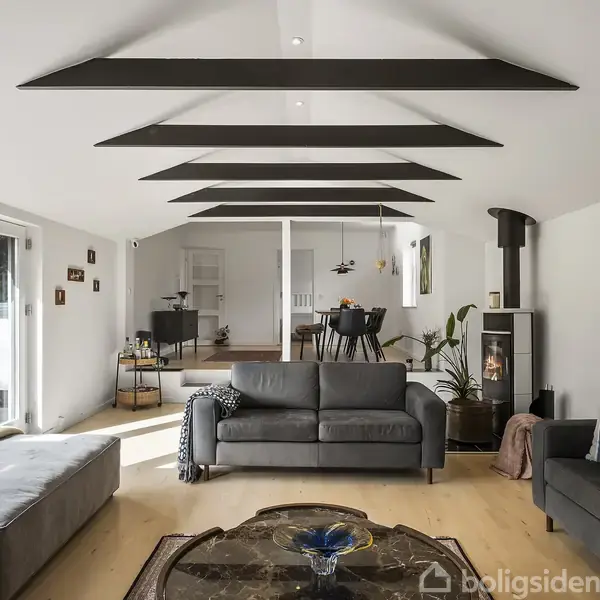 En moderne stue med grå sofaer står centralt, flankerede af en pejs til højre. Rummets lyse trægulv og store vinduer skaber en lys atmosfære. I baggrunden ses spiseområdet.