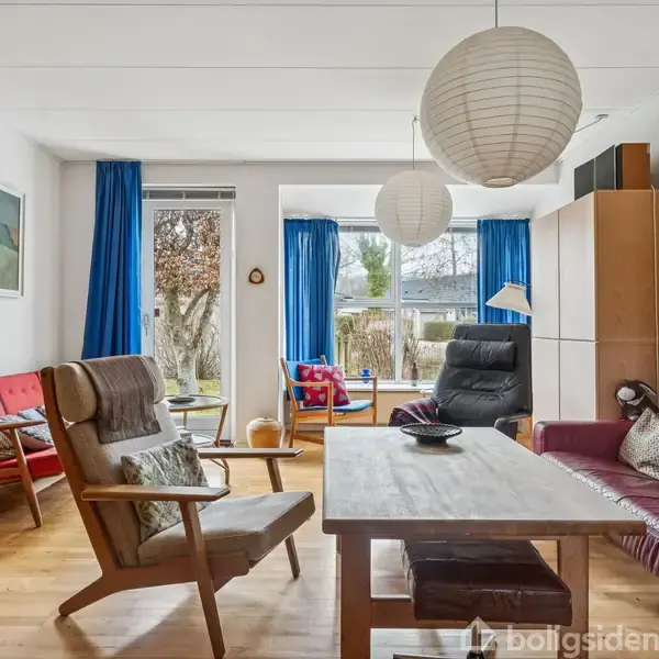 Et lyst stueområde med træmøbler, herunder en træstol og et sofabord. En rød sofa står til højre. Store vinduer med blå gardiner giver udsigt til haven udenfor.