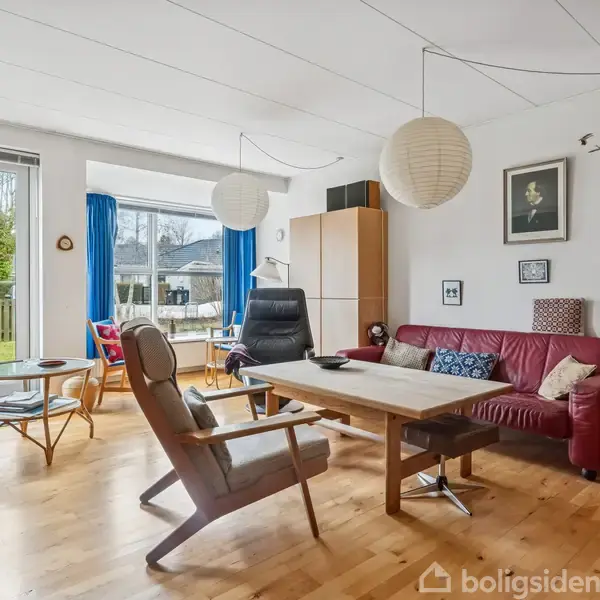 Stue med rødt lædersofaarrangement, lænestol og træbord. Store vinduer med blå gardiner fører ud til en have. Indretningen inkluderer lamper, billeder og en dansk flagdekoration.