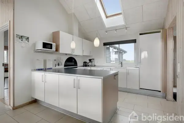 Et moderne køkken med hvidt interiør og store skabe. Et køleskab står til højre, en mikrobølgeovn hviler over et skab. Ovenlys og vindue giver naturligt lys. Ingen tekst.