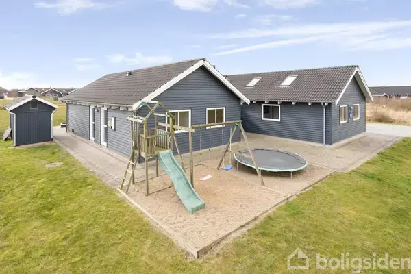 Et gråt hus ligger i en have med en legeborg med rutsjebane og en rund trampolin. Bygningen har mørkt tag og et skur i baggrunden.