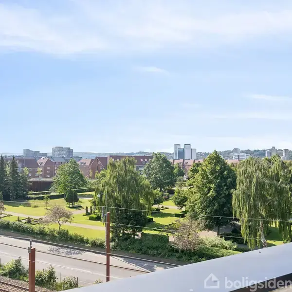 En grøn park med træer og buske beliggende nær en jernbane i bymiljø, omgivet af bygninger og himmel med få skyer.