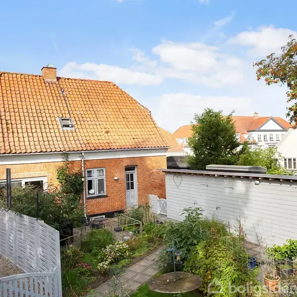 Et rødt murstenshus med rødt tegltag står i en have med stier og grønne planter. I baggrunden ses flere lignende huse under en blå himmel med skyer.