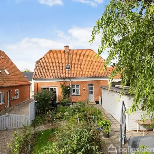 Et rødt murstenshus står i en have med grønne planter. Haven er omgivet af en hvid låge, og flisebelagte stier fører til huset. To sidebygninger ses også.