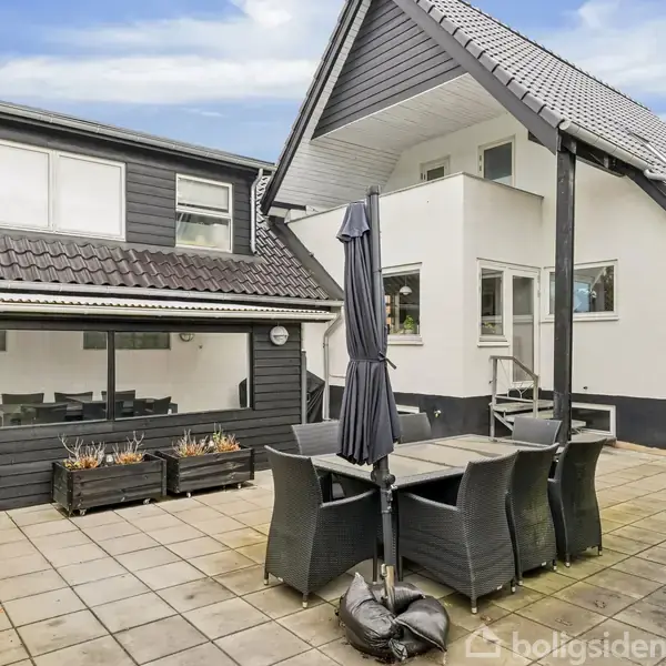 En have med flisebelagt terrasse, hvor sorte havemøbler med en parasol står samlet. Omgivet af en moderne, sort-hvid bygning med store vinduer og en velplejet hæk.
