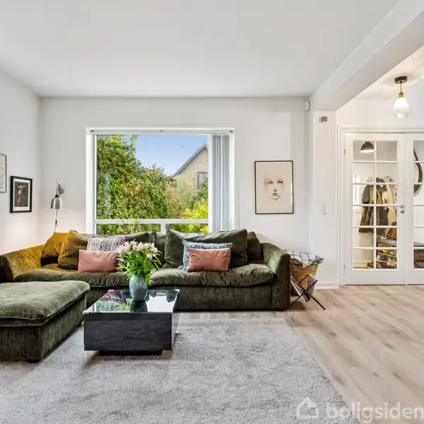 Grøn sofa står i en lys stue med et stort vindue, der viser en have udenfor. Et sofabord med en vase fyldt med blomster står foran sofaen. Væggene har kunstværker.