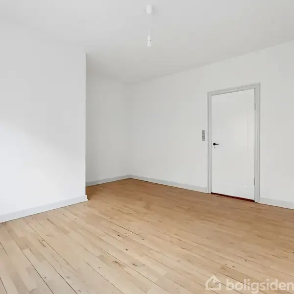 Et tomt rum med hvidmalede vægge og et lyst trægulv. En lukket dør er synlig i baggrunden. Rummet er enkelt og minimalistisk.
