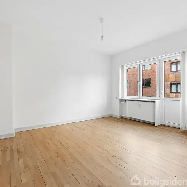 Et tomt rum med trægulv har et stort vindue og en dør mod højre, der fører ud til en balkon eller terrasse. Væggene er hvide, og lyset strømmer ind.