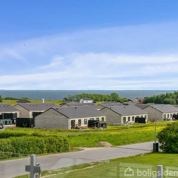 Åben stue og køkken med lyse trægulve. To barstole står langs køkkenøen. Rundt spisebord med seks stole i midten. Store vinduer giver lys. Vægmonteret fladskærm i baggrunden.